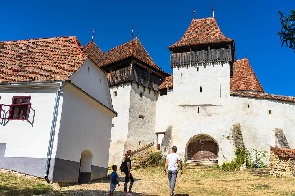 Family walking toward the entrance of Viscri historic fortified church with defensive towers and red tile roofs.